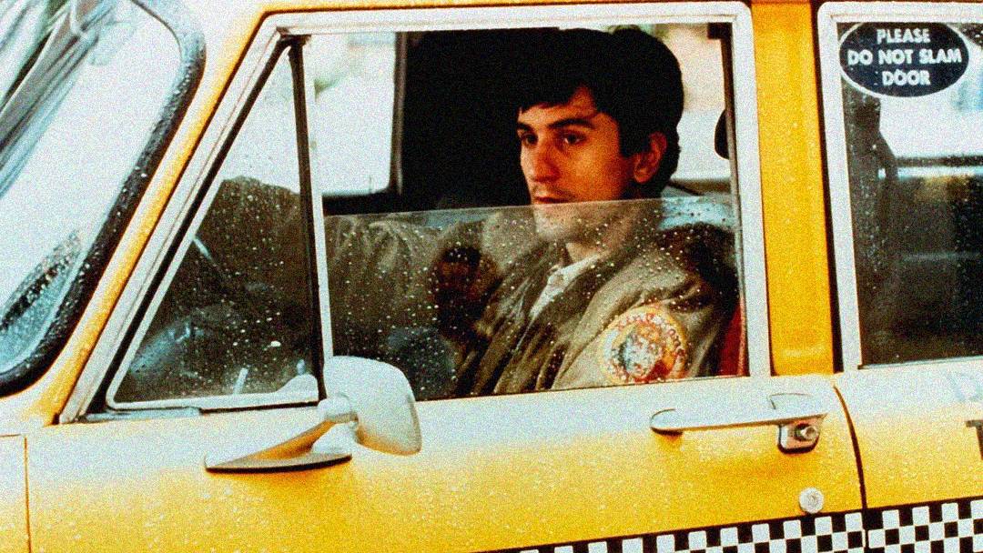 A man wearing a jacket sits behind the wheel of a yellow taxi cab, looking out the window. The window is partly rolled down, and a sign on the door reads "PLEASE DO NOT SLAM DOOR.