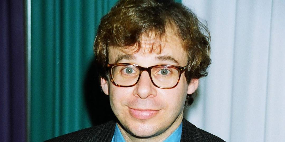 A man with short, wavy brown hair and glasses smiles slightly at the camera. He is wearing a blue shirt and a dark jacket, standing in front of a backdrop with teal and white curtains.