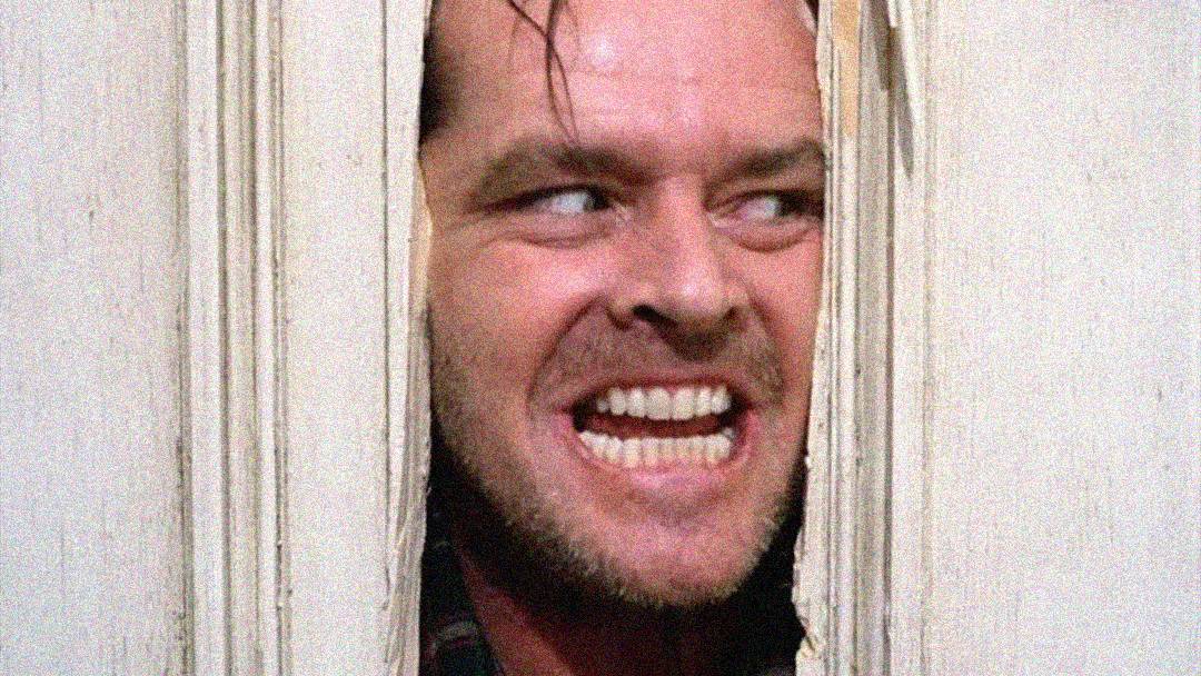 A man with a wild expression and bared teeth peers through a broken door, his face partially framed by jagged wood.