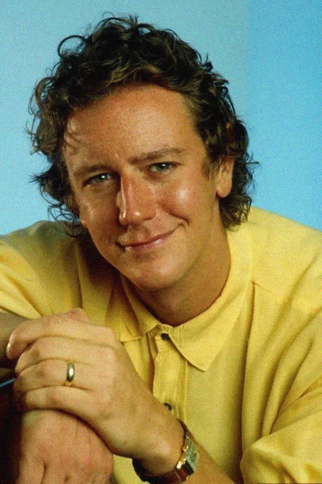 A person with curly brown hair and blue eyes, wearing a yellow collared shirt and a wristwatch, smiles gently while resting their hands together. The background is a soft blue.