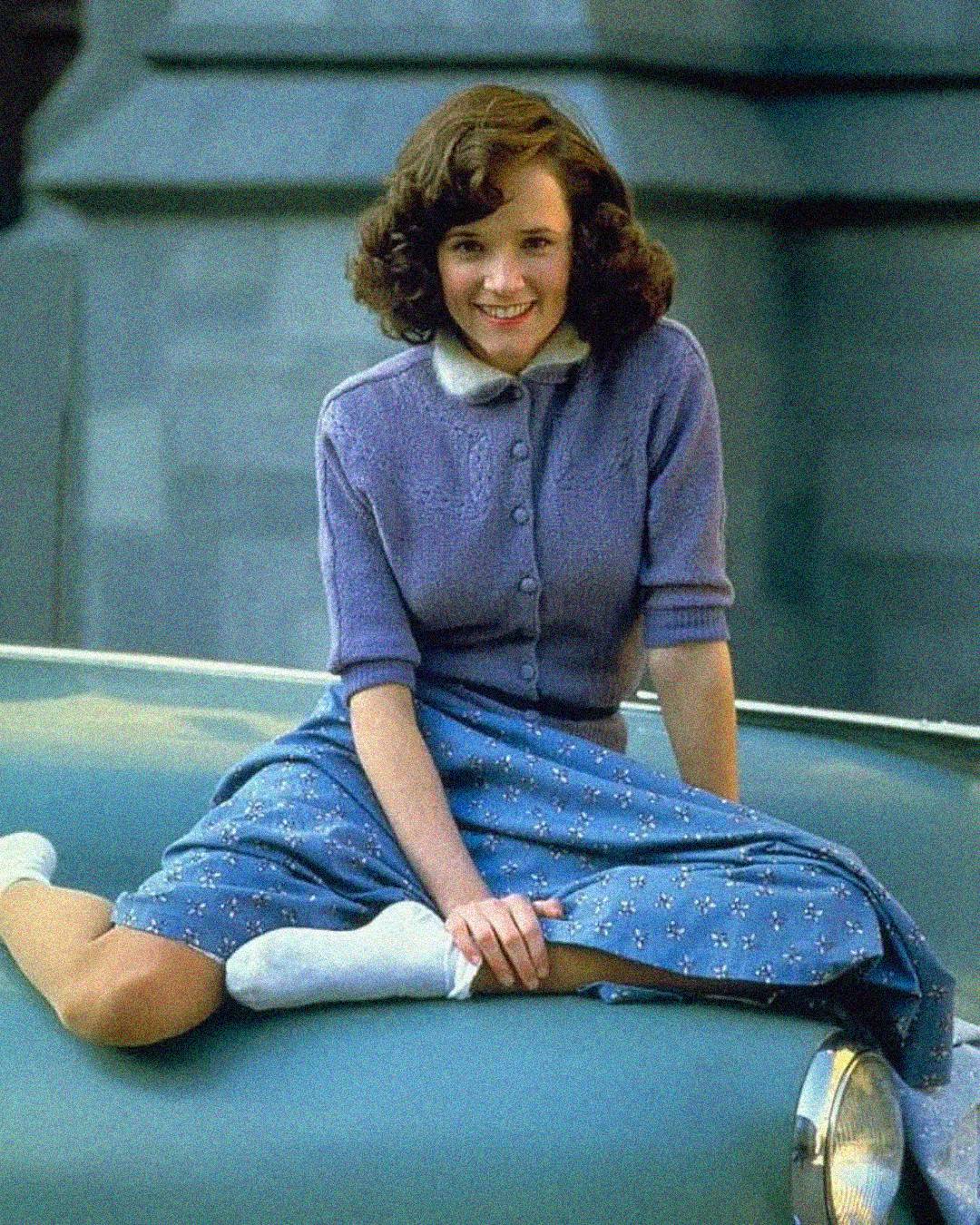 A young woman with wavy brown hair, wearing a lavender cardigan, blue floral skirt, white socks, and smiling, sits cross-legged on the hood of a light blue vintage car.