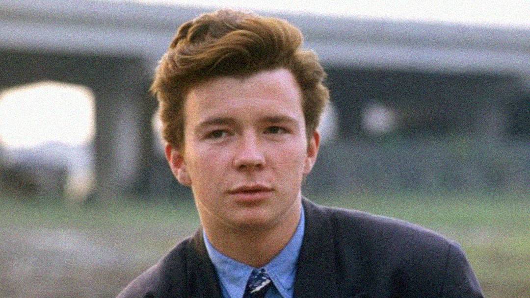 A young man with styled brown hair wears a suit and patterned tie, looking directly at the camera outdoors with a blurred overpass in the background.