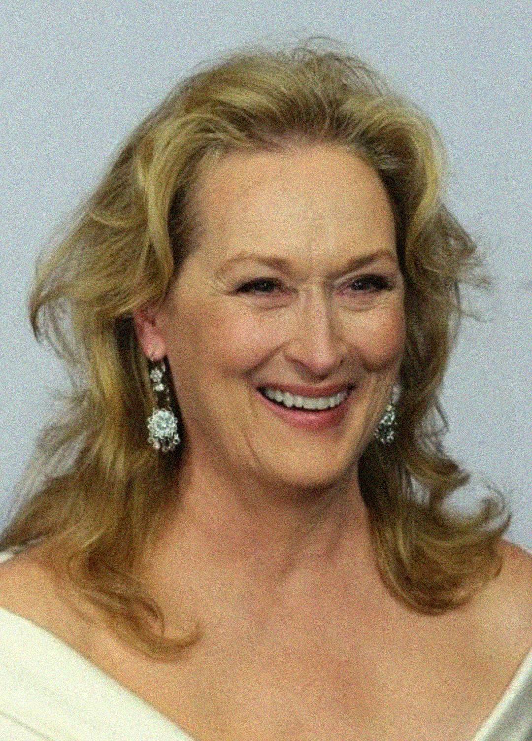 A woman with blonde wavy hair, wearing dangling silver earrings, a white off-shoulder outfit, and smiling against a plain light background.