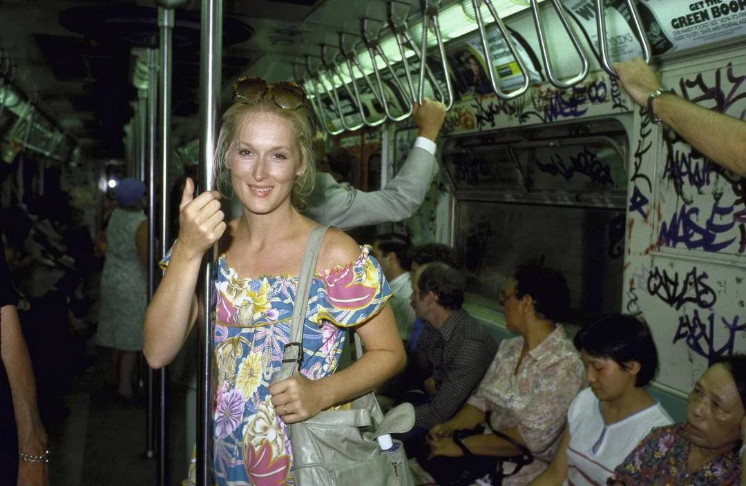 A woman in a floral dress stands and smiles on a graffiti-covered subway train, holding onto a pole. Other passengers sit or stand around her, and the train is busy and brightly lit.