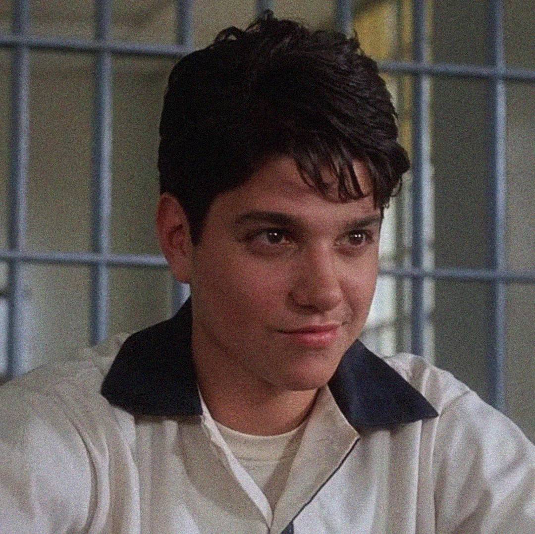 A young man with dark hair and a white shirt with a dark collar sits in front of metal bars, possibly in a jail or detention setting, looking slightly to the side with a subtle smile.