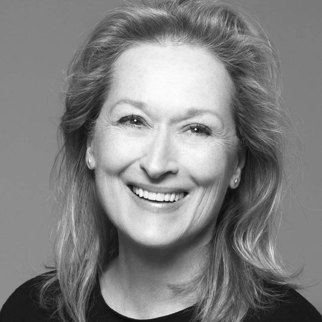 A woman with light hair smiles warmly at the camera in this black and white portrait. She is wearing a simple top and small stud earrings, with her hair softly styled away from her face against a plain background.