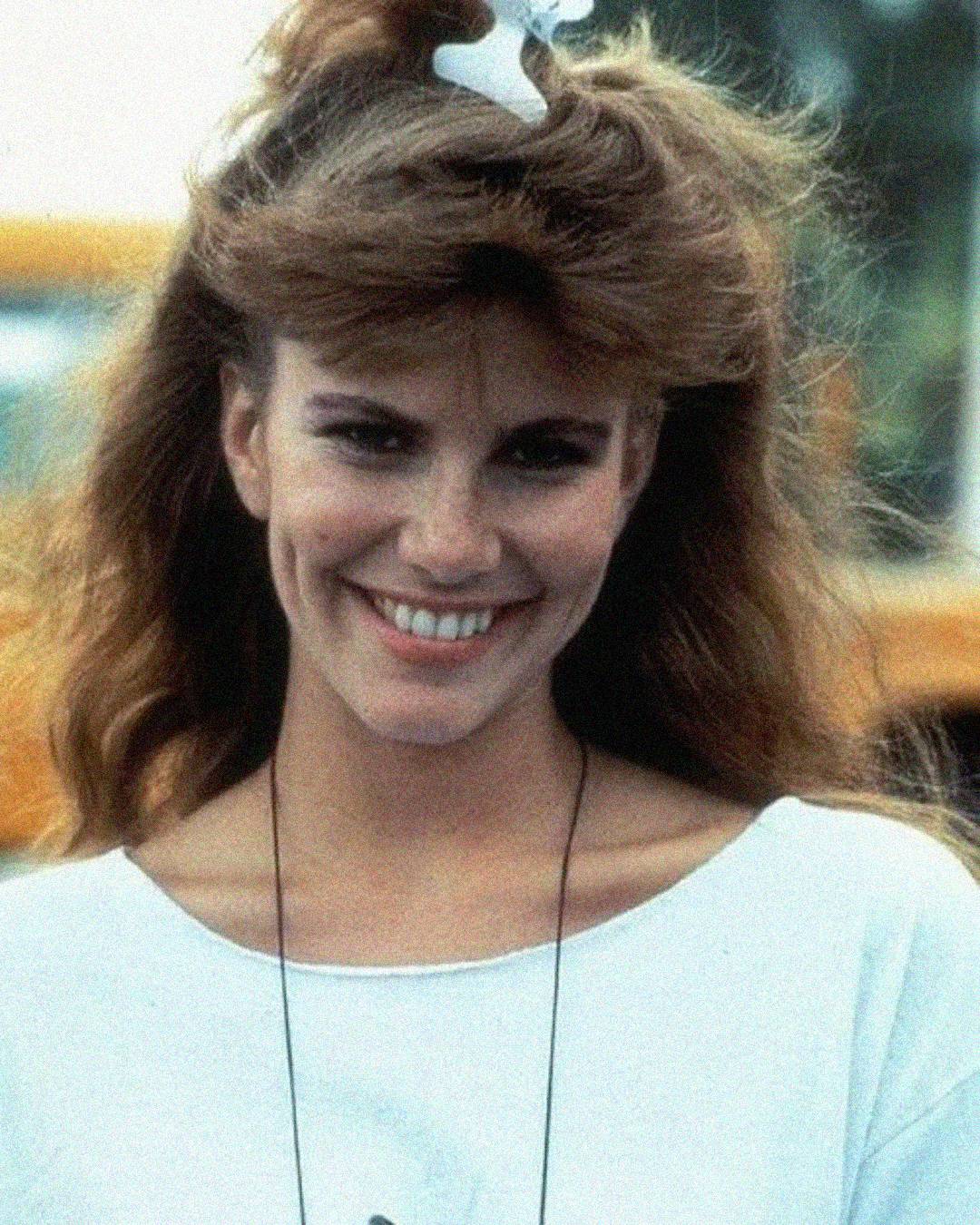A woman with long brown hair, styled with a white hair clip, smiles outdoors. She is wearing a white top and necklace, and the background appears to be sunny with blurred yellow shapes behind her.