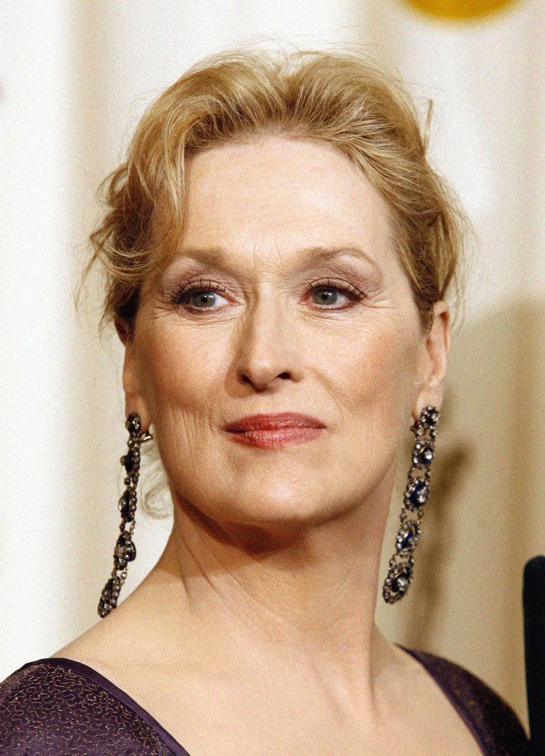 A woman with light skin, blonde hair styled up, and wearing long, dangling earrings looks slightly to the side with a composed expression, dressed in a dark top against a light background.