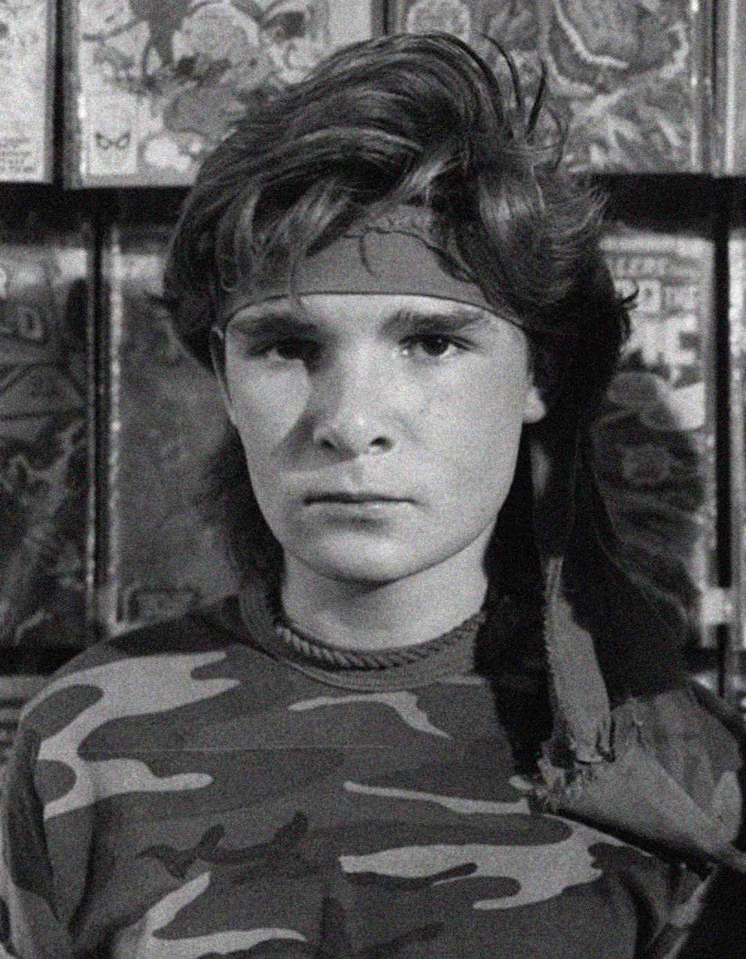 A young person with wavy hair wears a headband and camouflage shirt, standing in front of a display of comic books. The image is in black and white.