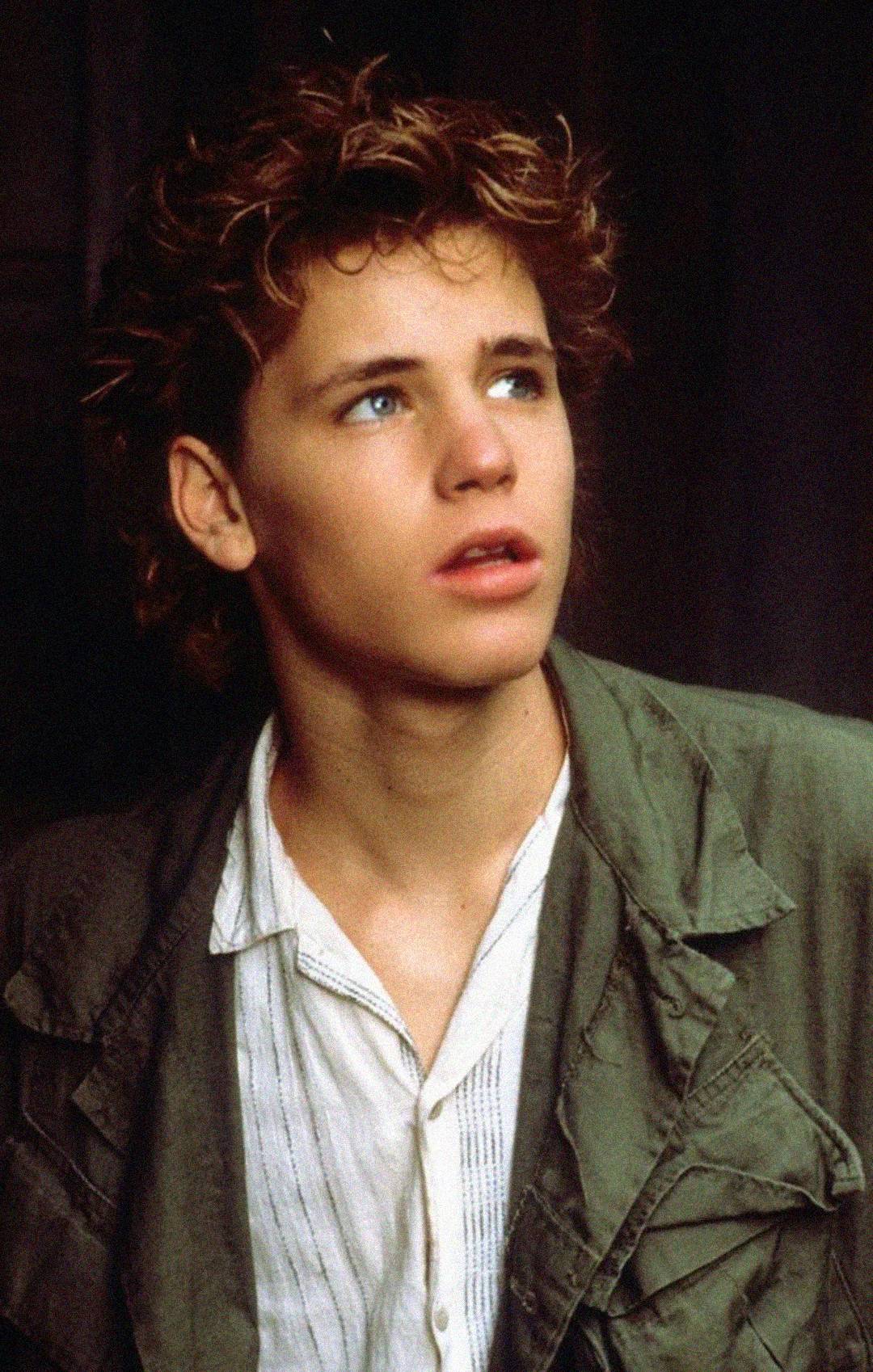 A young person with wavy, light brown hair looks up and to the side with a thoughtful expression. They are wearing a collared white shirt under a green jacket. The background is dark and out of focus.