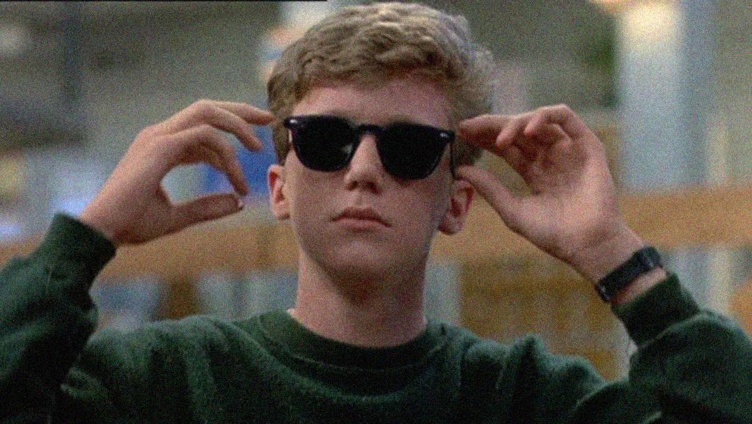 A young person with short, light brown hair and a green sweater puts on dark sunglasses, looking confident. The background is blurred, with soft lighting and indistinct indoor surroundings.