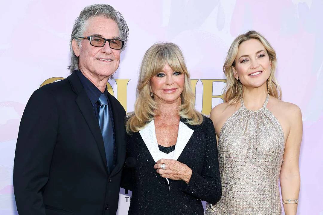 Three adults stand closely together, smiling at the camera. The man on the left wears glasses, a dark suit, and a tie. The two women wear elegant dresses, one in black and one in a shimmering silver gown, against a light background.