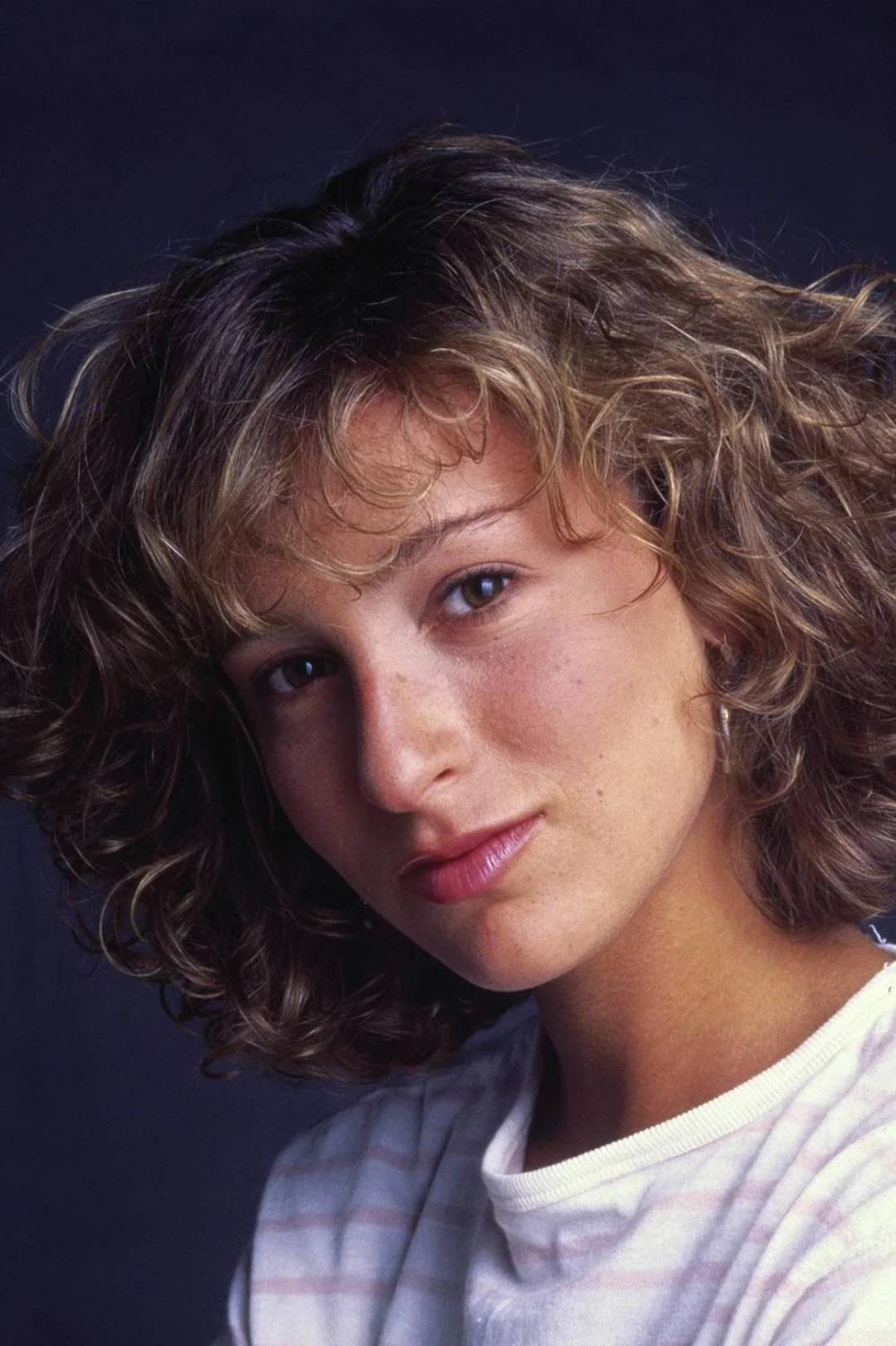 A young woman with curly, light brown hair and a striped shirt looks at the camera against a dark background. She has a neutral expression and soft natural makeup.