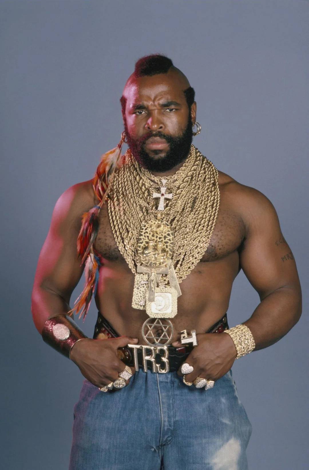 A muscular man with a mohawk wears many gold chains, bracelets, and rings. He is shirtless, wearing blue jeans, with a serious expression, standing against a plain gray background.