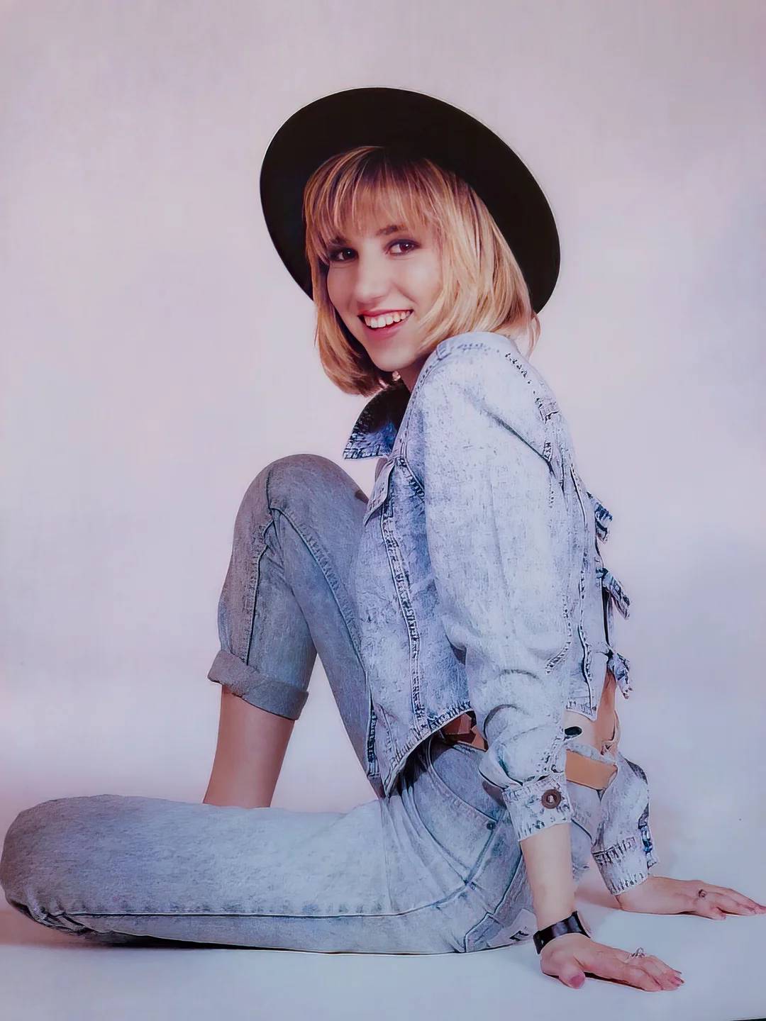 A woman with blonde hair wearing a black hat, light denim jacket, and matching jeans sits on the floor, smiling at the camera against a plain background.