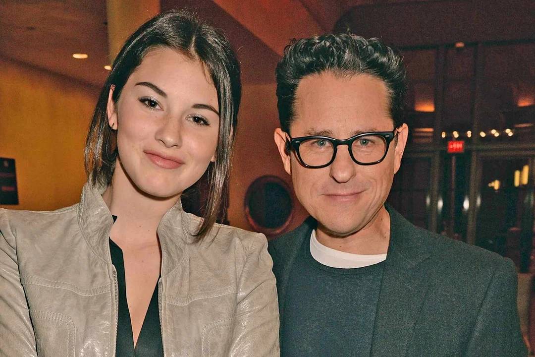 A young woman with straight brown hair stands next to a man wearing glasses and a dark blazer. Both are smiling slightly and posing together indoors with warm lighting in the background.