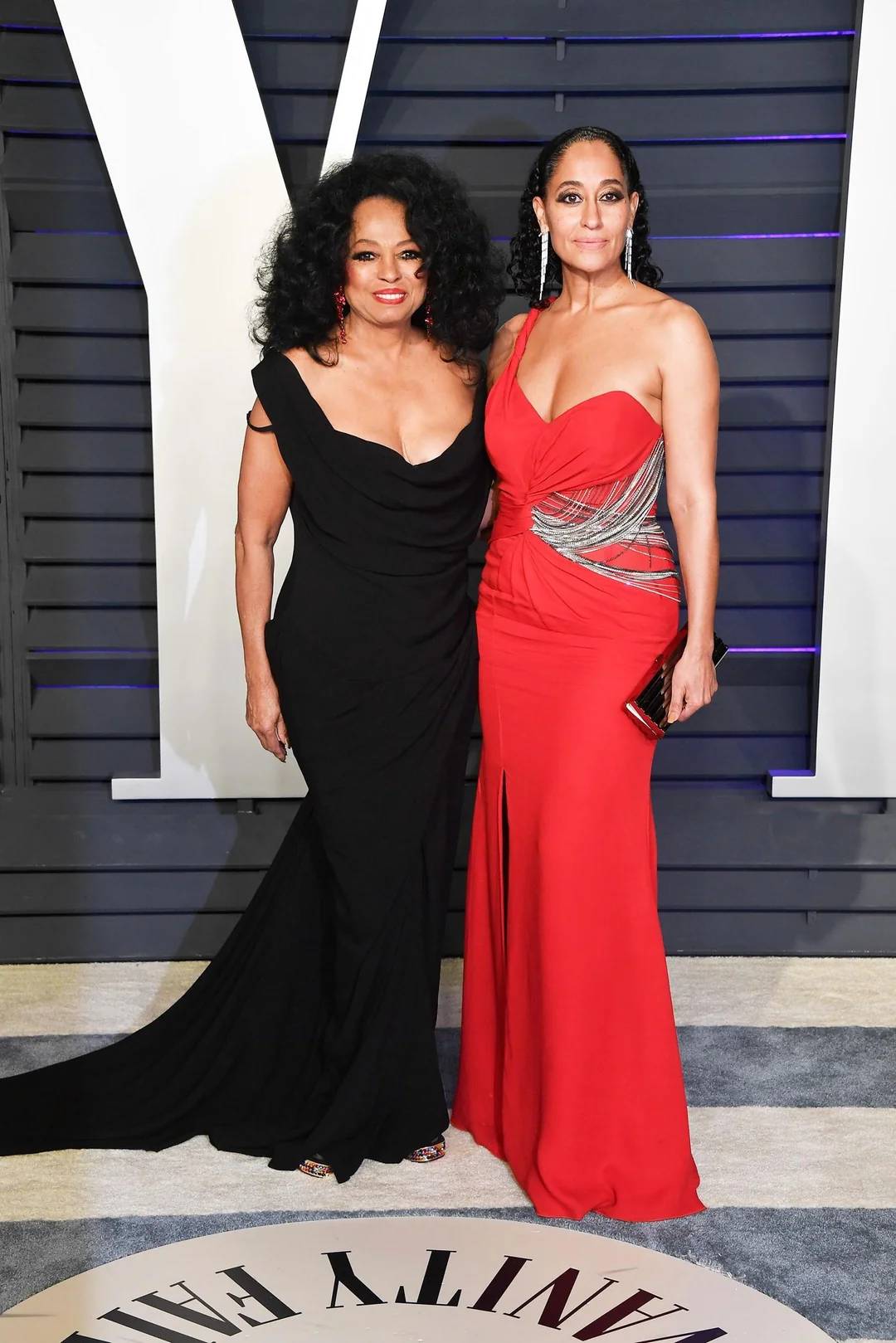 Two women pose together at an event; one wears a black off-the-shoulder gown with voluminous hair, the other wears a red strapless gown with silver accents and braided hair. They stand on a carpet with "Vanity Fair" written on it.