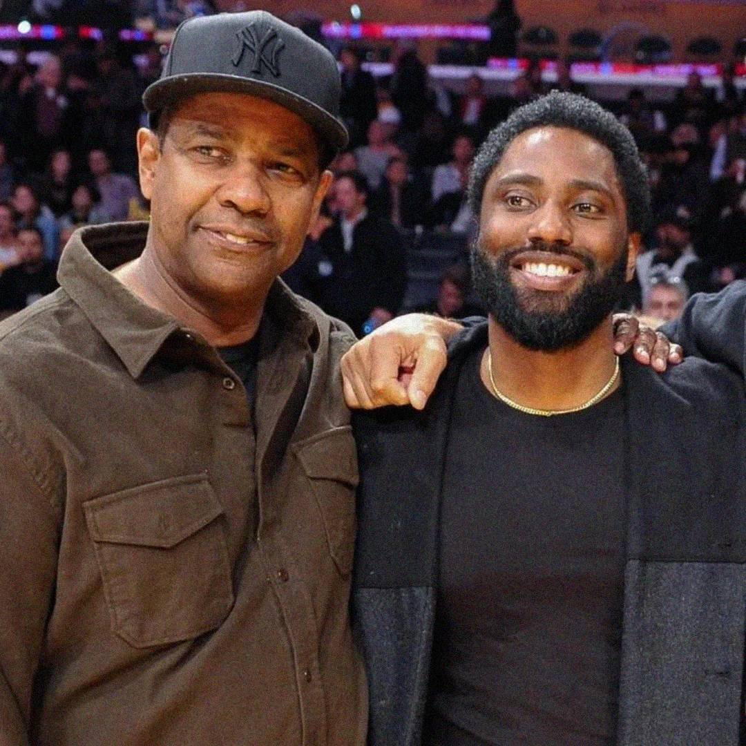 Two men are smiling and posing together at an indoor event. One is wearing a brown button-up shirt and a black baseball cap; the other has a beard and is wearing a black shirt and coat. People are seated in the background.