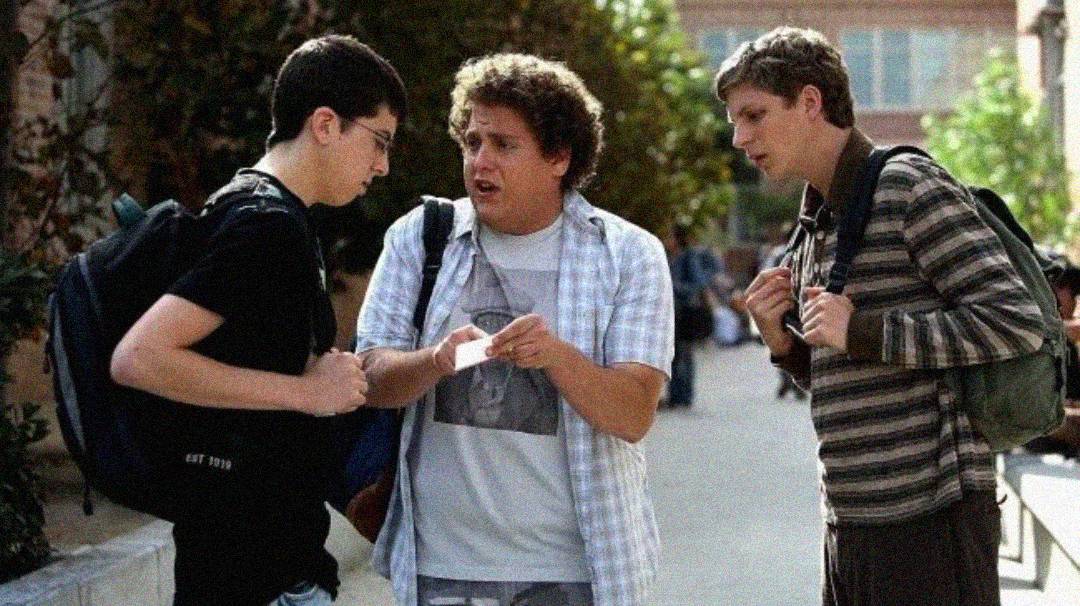 Three young men with backpacks stand outside, talking closely together. One points at an object in another's hand, while the third person looks on attentively, all appearing to be engaged in a serious conversation.