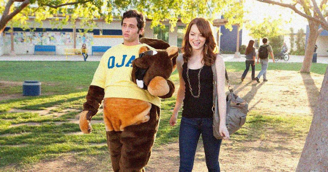 A young woman walks outside on a sunny day beside a person wearing a brown bull mascot costume and a yellow shirt that says "OJA." Trees and several people are visible in the background.