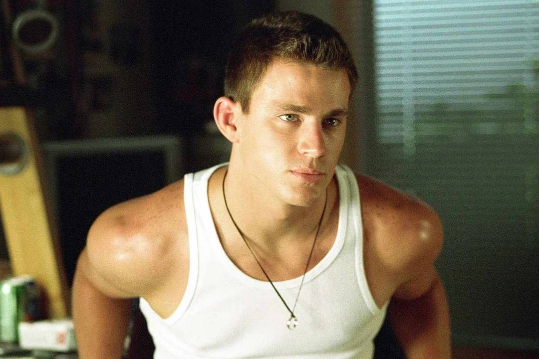 A young man with short brown hair wearing a white tank top and a necklace sits indoors, looking slightly to the side with a serious expression. The background is dimly lit, with some objects and a window visible.