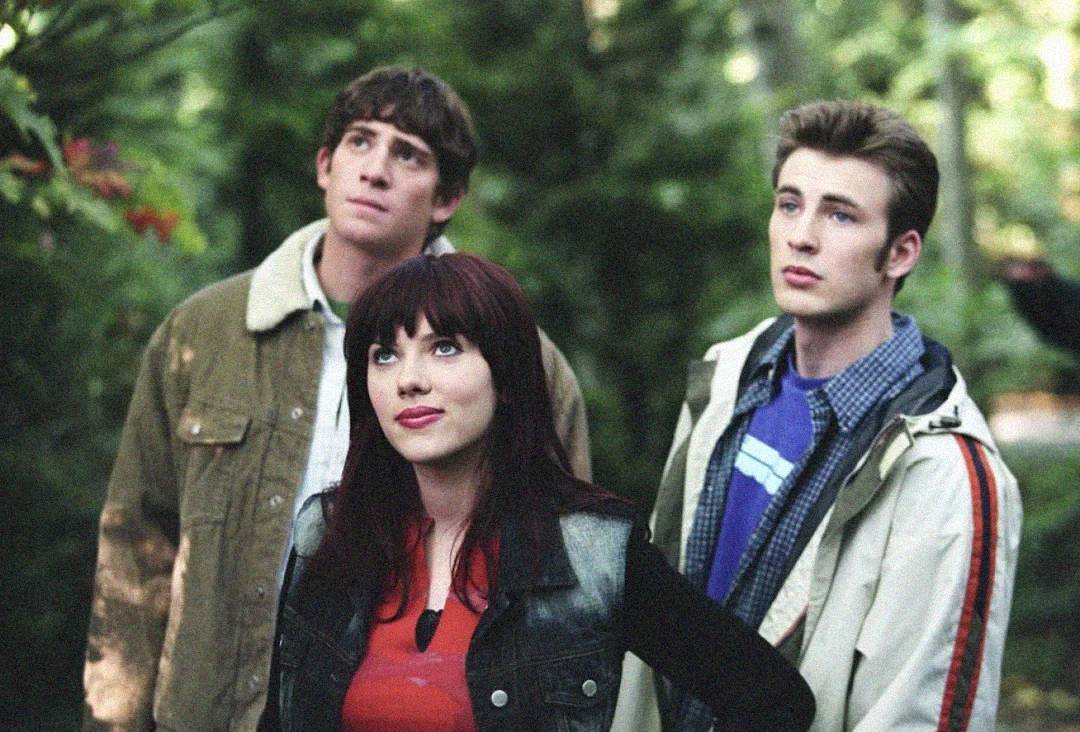 Three young adults stand outdoors in a wooded area, looking slightly upward. The woman with dark hair is in front, flanked by two men with short hair, all dressed in casual jackets. Lush greenery surrounds them.