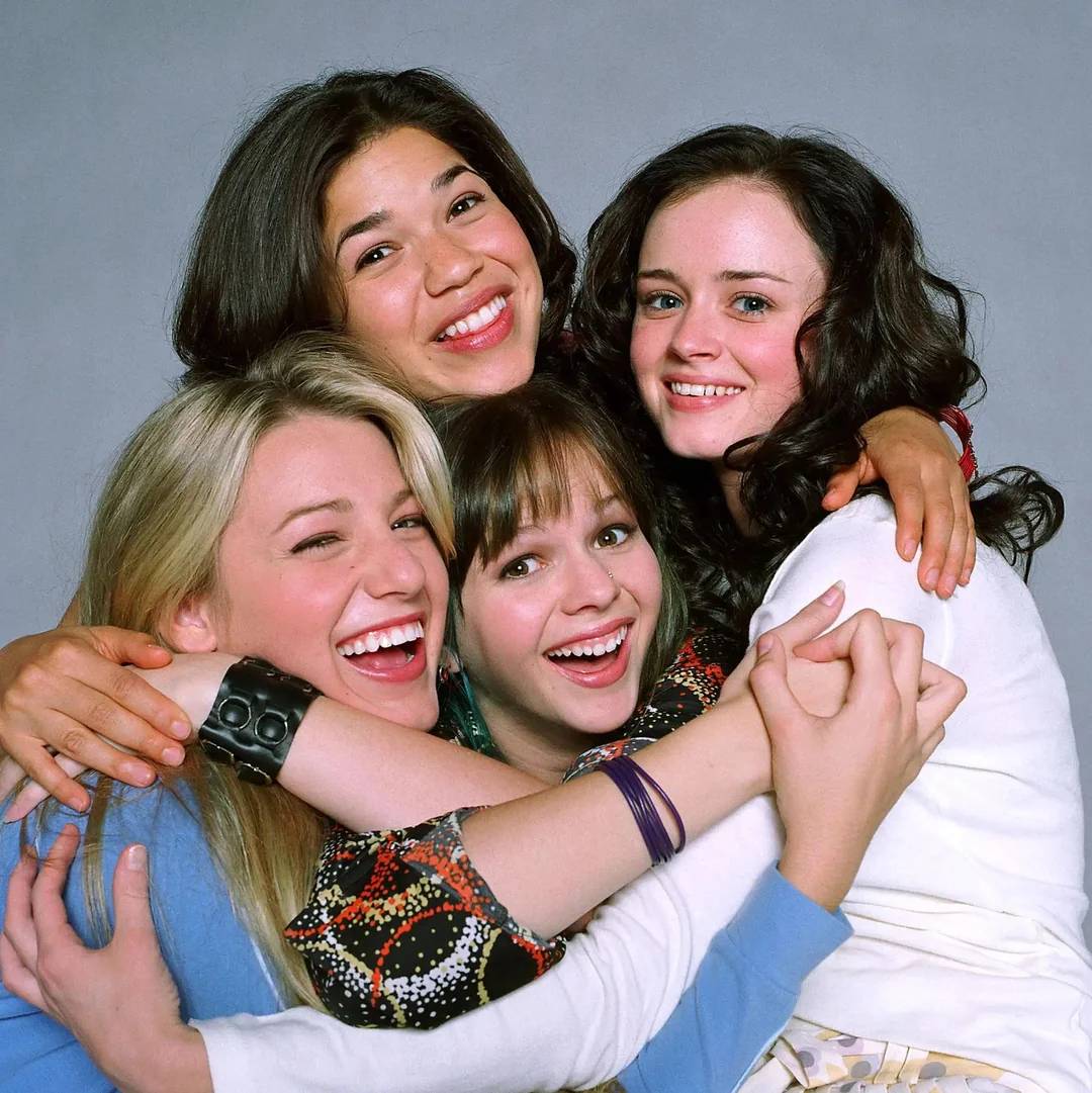 Four young women smile and hug each other closely in front of a plain background, showing a joyful and friendly moment.