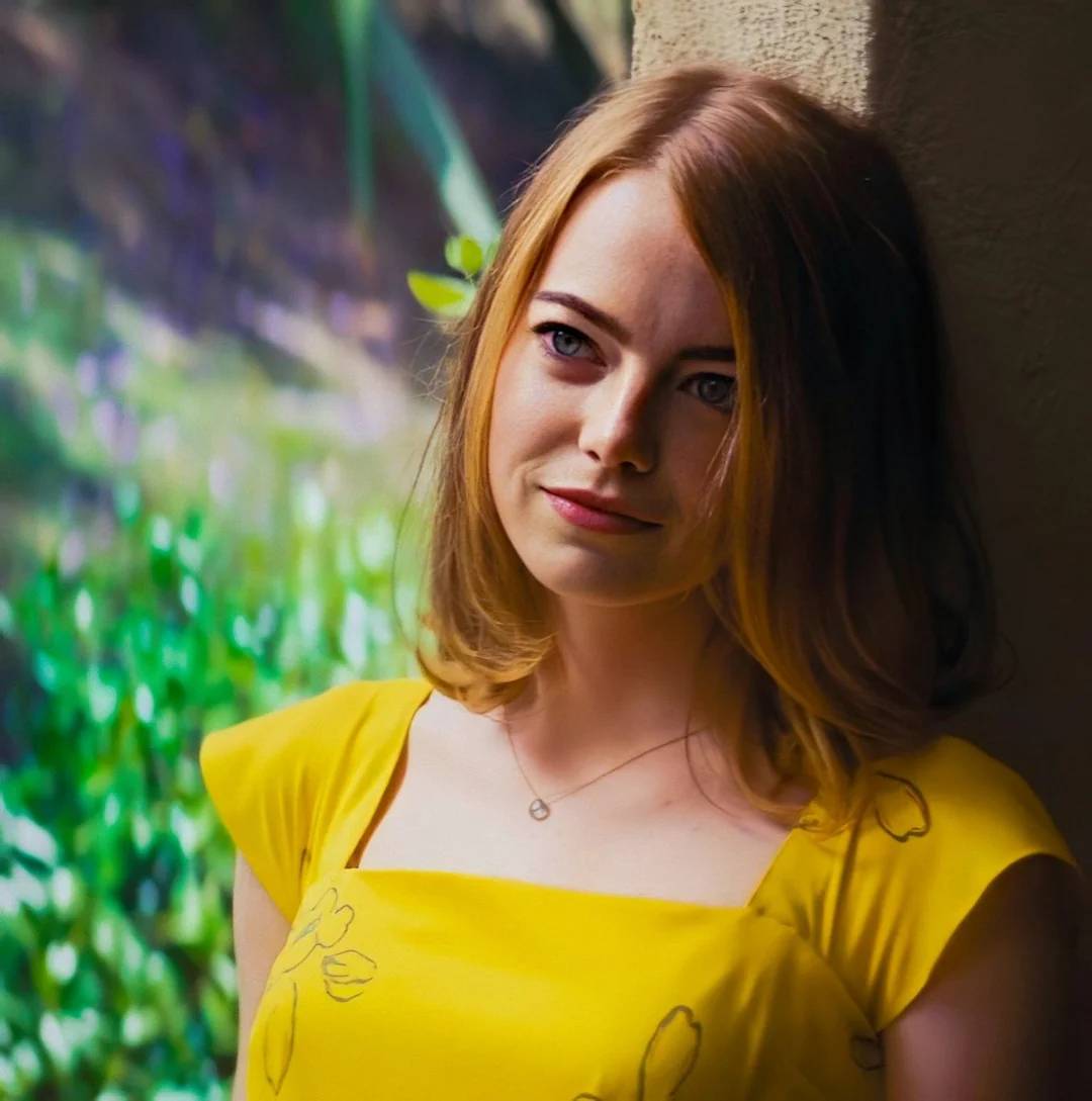 A woman with light skin and straight, shoulder-length blonde hair leans against a wall, wearing a bright yellow dress. The background is blurred with green foliage and sunlight.