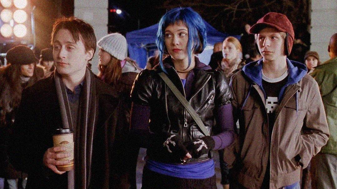 Three young adults stand together outdoors at night, surrounded by people. The center person has bright blue hair and wears a black jacket, flanked by two others in casual clothes; one holds a coffee cup, the other wears a red cap.