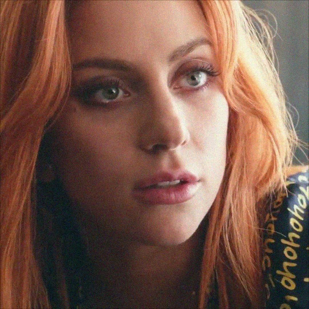 A woman with long, wavy, orange hair and light eyes looks slightly to the side. She has a thoughtful expression and is wearing a dark top with yellow writing on the shoulder.