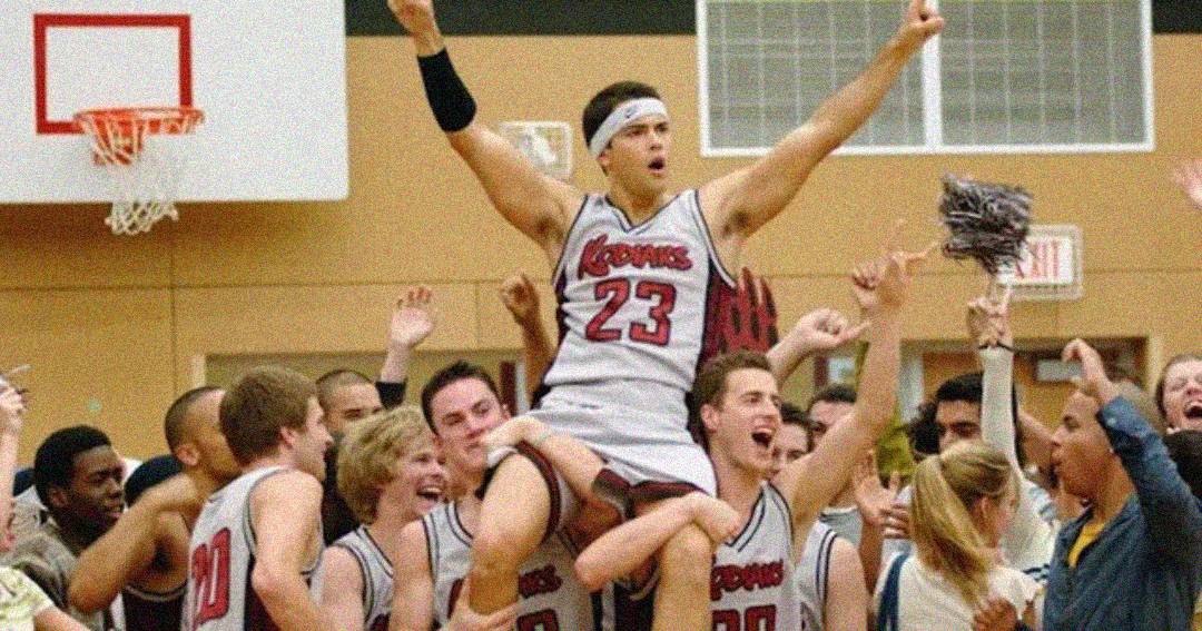 A basketball player wearing a number 23 jersey is lifted by cheering teammates and fans on a court, celebrating a victory. The player raises one arm triumphantly.