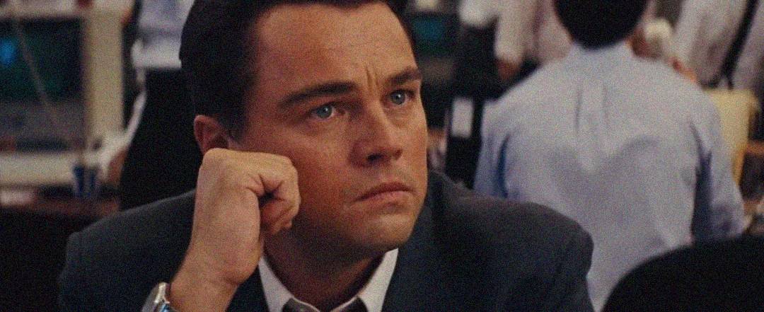 A man in a suit sits at a desk with his hand resting on his face, looking serious and deep in thought. People and computer monitors are visible in the blurred background, suggesting a busy office setting.
