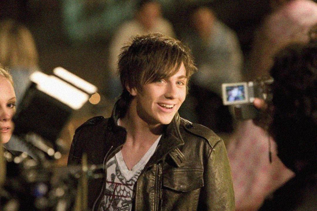 A young man with shaggy brown hair smiles while wearing a leather jacket. He is surrounded by people and cameras, suggesting he is being filmed or interviewed. The background is blurred with soft lighting.
