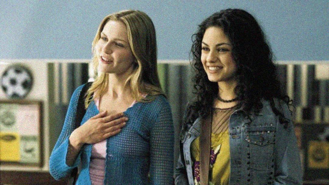 Two young women stand side by side indoors, smiling. The woman on the left has blonde hair and wears a blue sweater, while the woman on the right has curly dark hair and wears a jacket over a yellow top. A blurred background is visible.