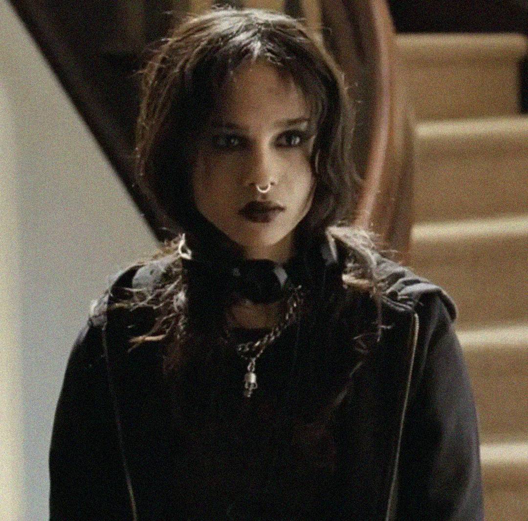A person with long dark hair, dark eye makeup, and black lipstick wearing a black hoodie and necklaces stands indoors in front of a staircase. The lighting is dim and the mood appears serious.