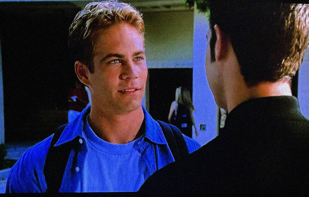 A young man with short blond hair, wearing a blue shirt and backpack, stands outside and talks to another person whose back is facing the camera. A blurred figure is visible in the background.
