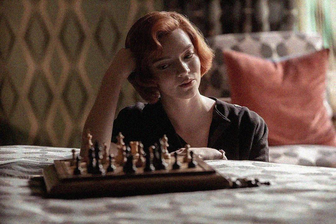 A woman with short red hair sits at a table, resting her head on her hand, thoughtfully looking at a chessboard arranged with pieces. The setting appears cozy with patterned curtains and a chair with a red cushion in the background.