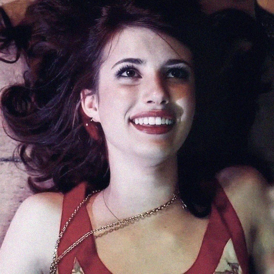 A young woman with long, dark wavy hair lies on her back, smiling brightly and looking upward. She is wearing a red top and a thin gold chain necklace. The lighting is soft, creating a warm and cheerful atmosphere.