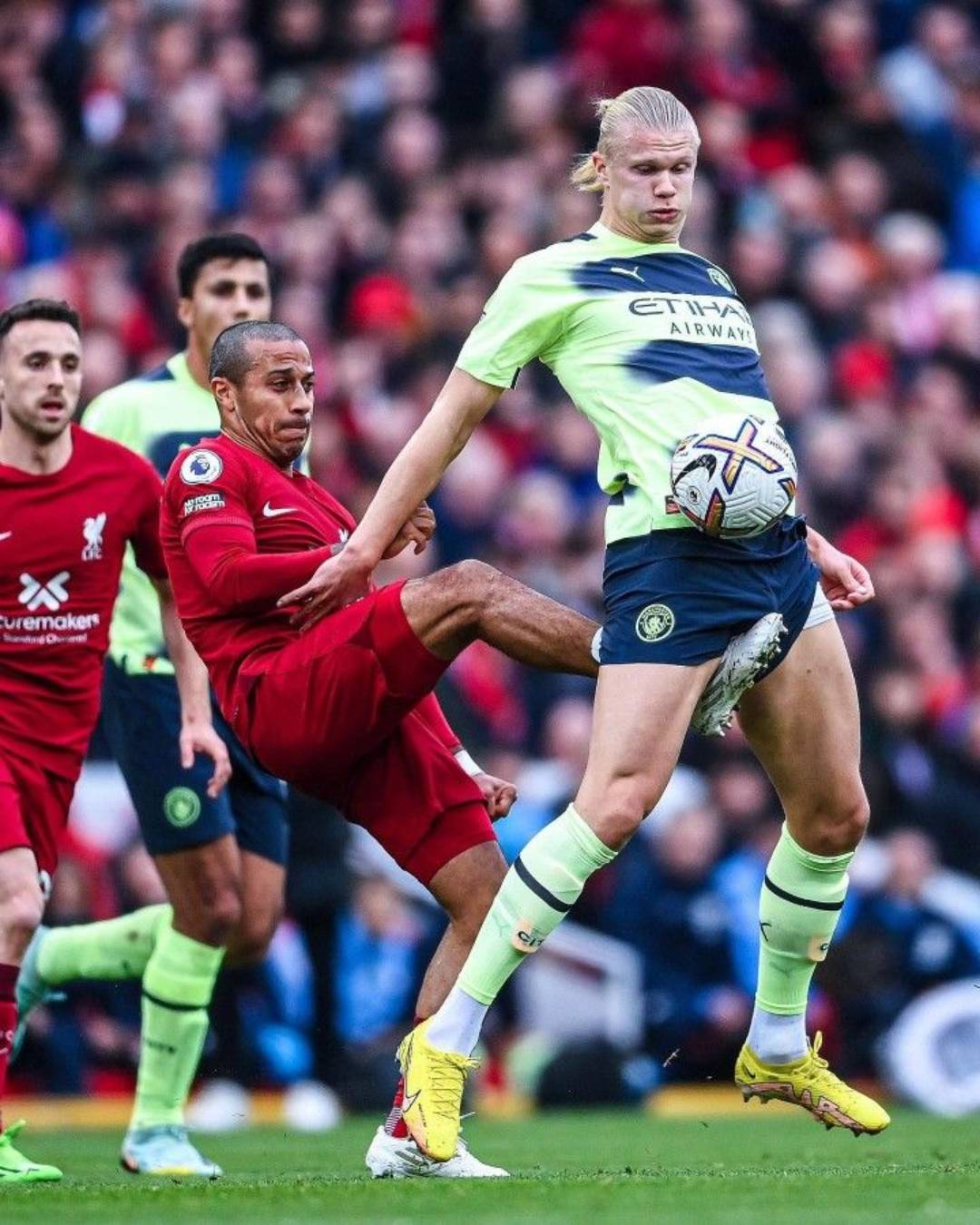A soccer player in a red kit attempts to kick the ball as a player in a light blue kit blocks it with his leg during a competitive match, with other players and a crowd in the background.