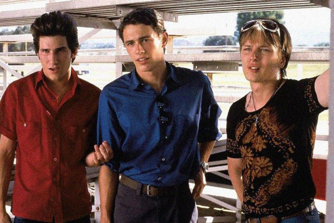 Three young men stand outdoors by metal bleachers. One wears a red shirt, one a blue shirt, and the third wears a printed black shirt with sunglasses on his head. They look in the same direction, appearing engaged or curious.