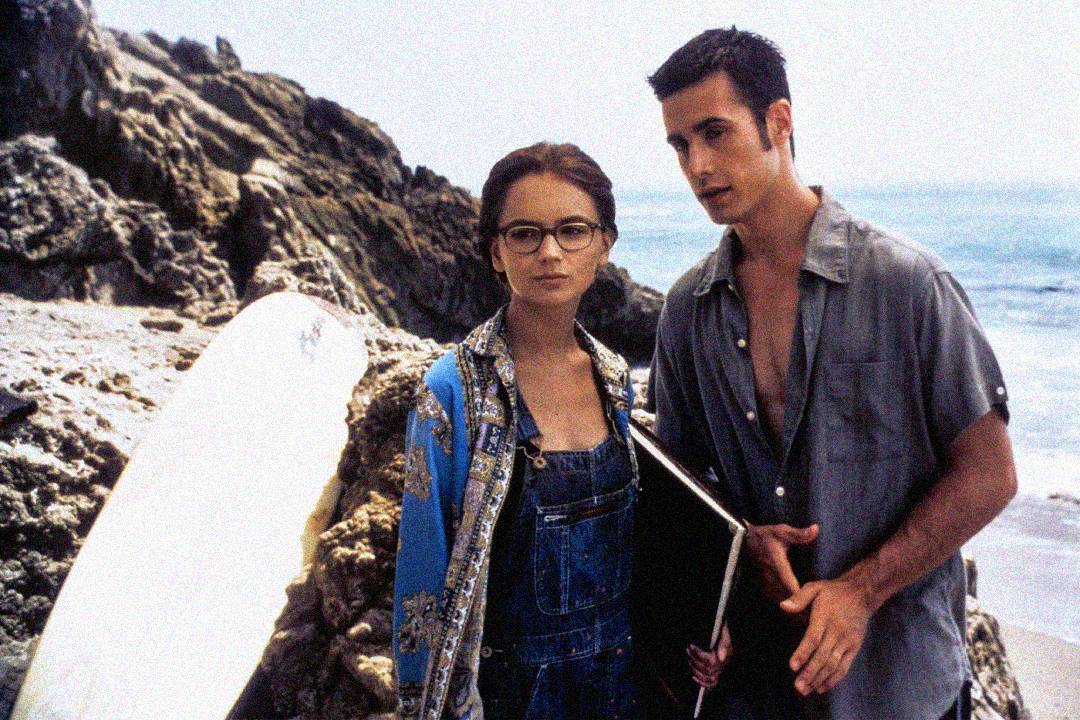 A young woman with glasses and a patterned jacket stands by rocky cliffs near the ocean, holding a folder. Beside her, a young man in an open shirt holds a surfboard, both looking ahead with serious expressions.