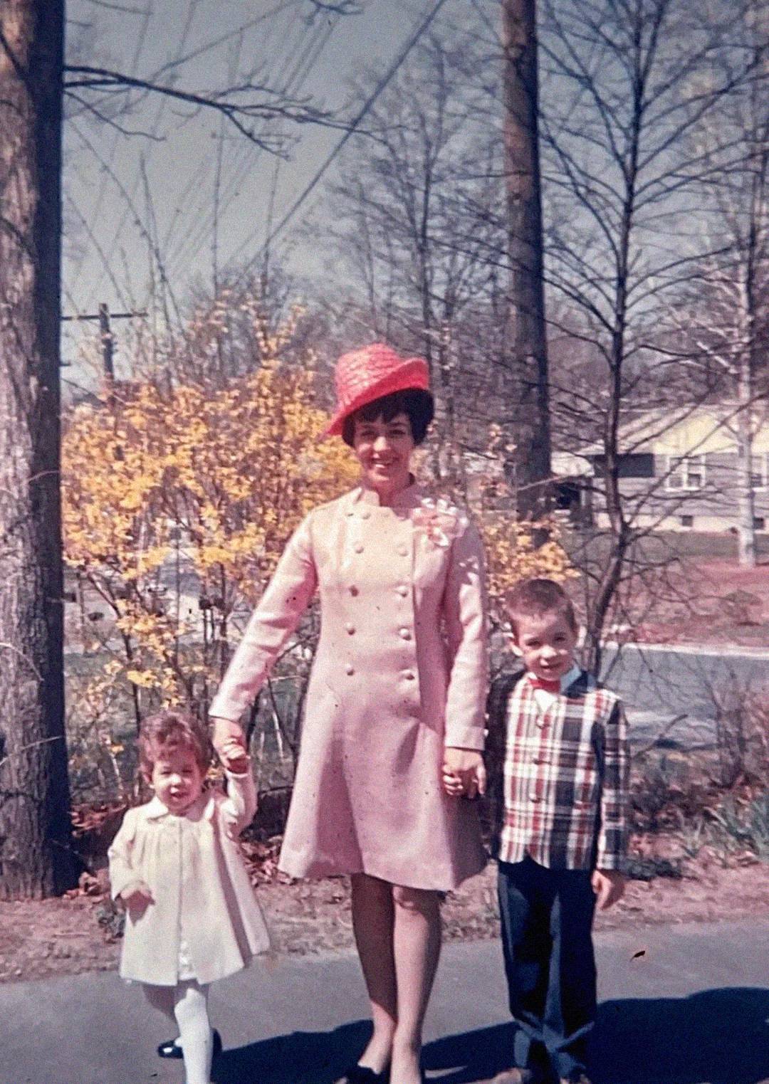 A woman in a pink coat and red hat holds hands with a young girl in a white dress and a boy in a plaid shirt, standing outside near trees and yellow blooming bushes on a sunny day.