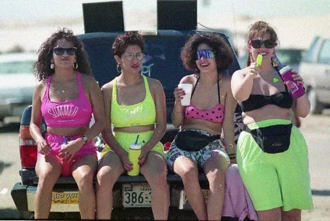 Four women in bright, retro summer outfits sit on the back of a vehicle at the beach. They wear sunglasses and fanny packs, holding drinks and water guns, smiling and enjoying a sunny day.