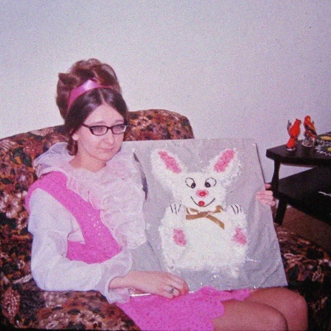 A woman wearing glasses and a pink dress with ruffles sits on a floral-patterned couch, holding a tray with a bunny-shaped cake decorated with a bowtie and pink ears. A small table with figurines is in the background.