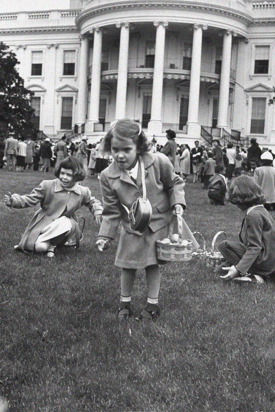 Black and white photo of young children searching for eggs on the lawn in front of the White House, carrying baskets, with adults and more children gathered in the background.
