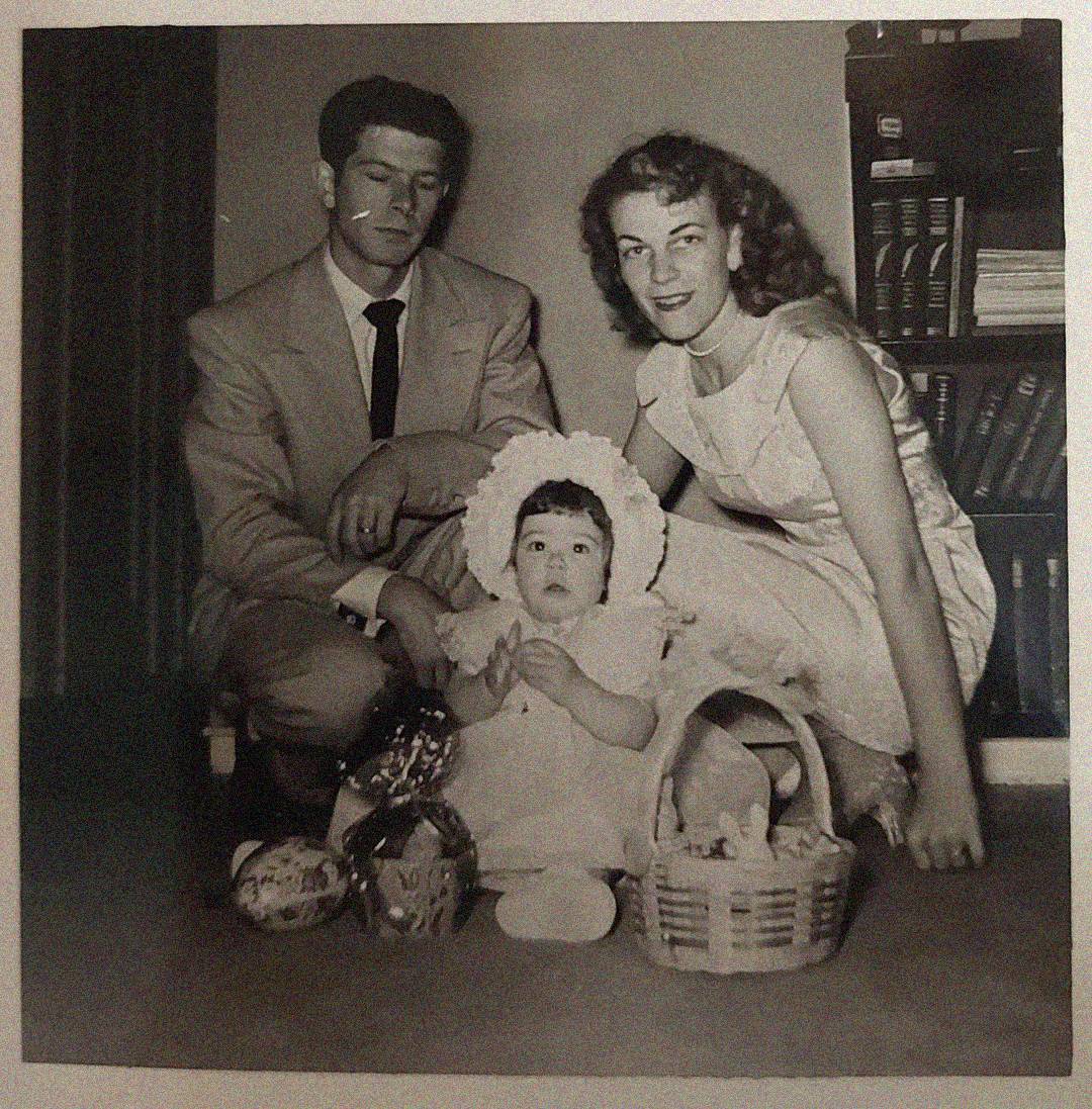 A vintage black-and-white photo of a man in a suit, a woman in a light dress, and a baby in a bonnet surrounded by baskets, seated together indoors in front of a bookshelf.
