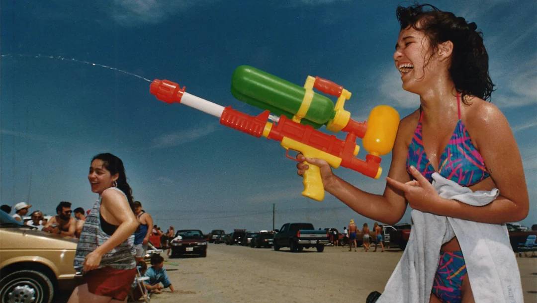 A smiling woman in a colorful swimsuit sprays a water gun at someone on a crowded beach parking lot, while others in swimwear enjoy the sunny day.