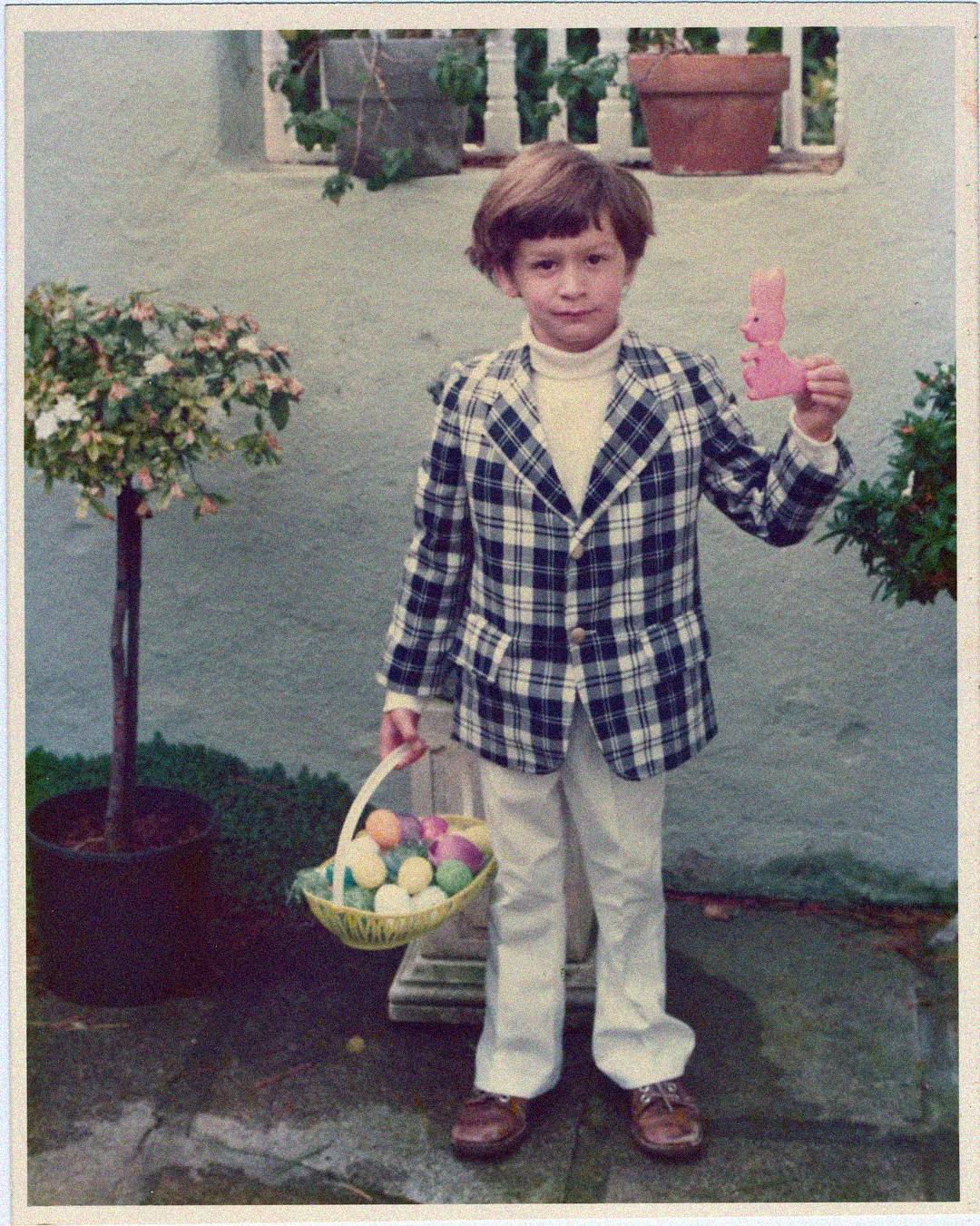 A young child wearing a plaid jacket, white pants, and brown shoes stands outdoors holding a basket of colorful Easter eggs in one hand and a pink bunny-shaped object in the other. Potted plants are in the background.