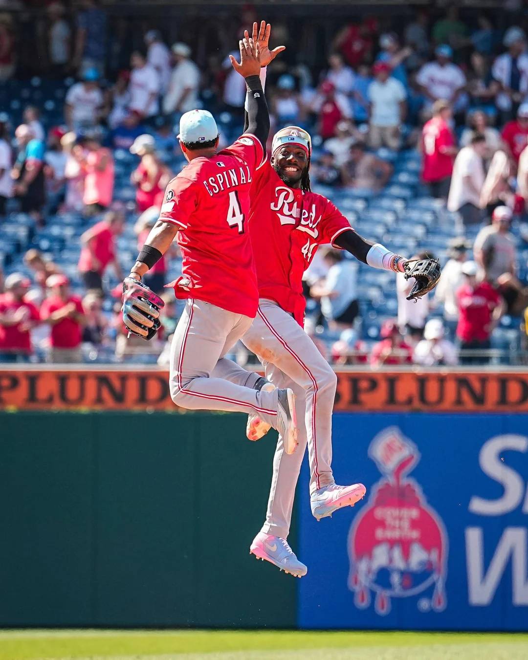 Two baseball players in red uniforms jump and high-five each other in celebration on the field, with fans watching from the stadium seats in the background.