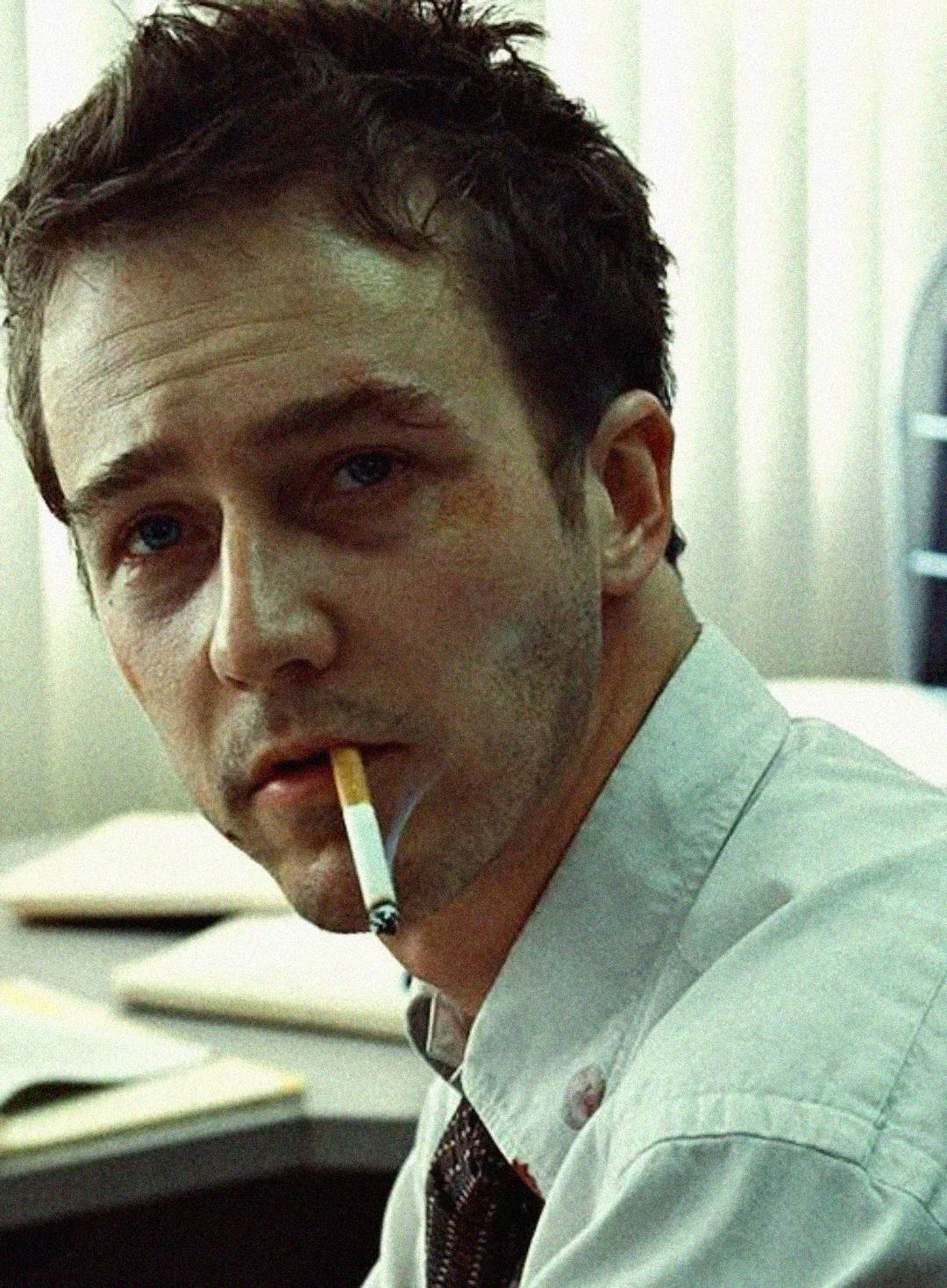 A man with tousled hair and a tired expression sits at a desk, wearing a white shirt and tie, with a cigarette hanging from his mouth. The background shows vertical blinds and office items.